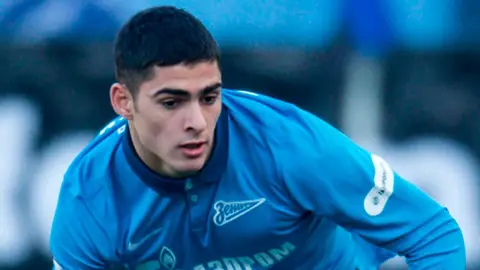 Ramil Şeydayev qışkı formaya bənzər geyimdə, top və stadion fonunda