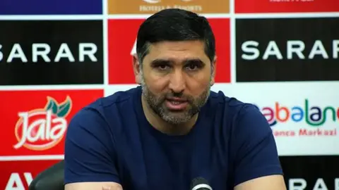 Sənan Qurbanov mikrofon qarşısında çıxış edir, fonunda stadion və U-15 oyunları